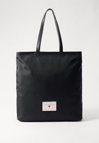 Black tote bag made of textured fabric, featuring two handles and a metallic logo plate at the bottom center. Spacious rectangular design.