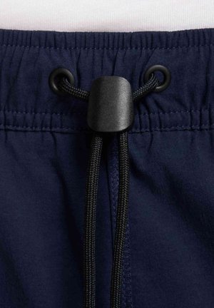 Ceinture élastique bleu marine avec cordon noir passant dans des œillets en métal noir, fixé par un verrouillage en plastique noir.