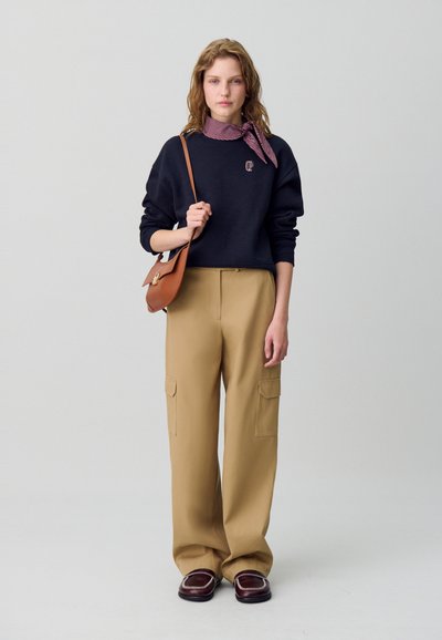 Sudadera de navy con logo, pantalones cargo beige con bolsillos laterales, bolso de cuero marrón y mocasines oscuros. Textura suave y corte relajado en general.