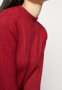 Rode trui met een hoge halslijn, gemaakt van zachte, gestructureerde stof. Heeft lange mouwen en een getailleerde pasvorm.