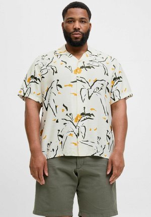 Camisa de manga corta en crema claro con patrones florales en negro y amarillo. Corte holgado, cuello con solapa y cierre de botones en el frente. Combinada con pantalones cortos verdes.