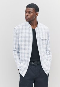 Camicia a quadri bianca e blu con bottoni, tessuto leggero, tasca singola sul petto, abbinata a una camicia nera e pantaloni scuri.