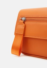 Bolsa de material sintético naranja con una textura suave, que cuenta con un bolsillo con cremallera y una correa texturizada en un color a juego.