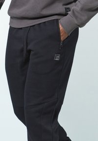 Schwarze Jogginghose aus weichem Stoff mit schmal zulaufendem Schnitt, ausgestattet mit einer Reißverschlusstasche und einem kleinen Logo-Patch an der Seite.