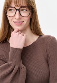 Vrouw met lang lichtbruin haar, tortoisebruine bril en een bruine, geribbelde trui, die zacht glimlacht met haar hand bij haar kin.