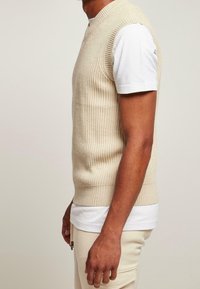 Beżowy dzianinowy bezrękawnik o prążkowanej strukturze, z dekoltem w kształcie litery V, noszony na białej koszulce, ukazany z boku.