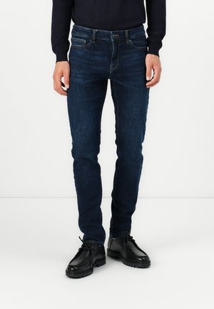 Mand iført mørkeblå slim-fit jeans, sort langærmet top og sorte læder snørestøvler, stående på hvid baggrund.