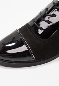 Chaussure habillée noire avec un bout en cuir verni brillant et des accents. Présente un dessus en daim avec une rangée de clous en argent décoratifs le long de la couture.
