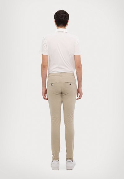 Polo shirt blanc à manches courtes pour homme associé à un pantalon beige slim-fit. Le pantalon est doté de deux poches arrière avec fermetures éclair. Des baskets complètent la tenue.