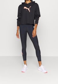 Cappuccio corto nero con logo Puma rosa, abbinato a leggings scuri a vita alta con accenti rosa e scarpe da ginnastica bianche.