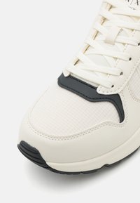 Sneaker blanche avec un mesh texturé et des matériaux synthétiques lisses, présentant des accents noirs contrastés et un design à lacets. Bout arrondi et semelle en caoutchouc.