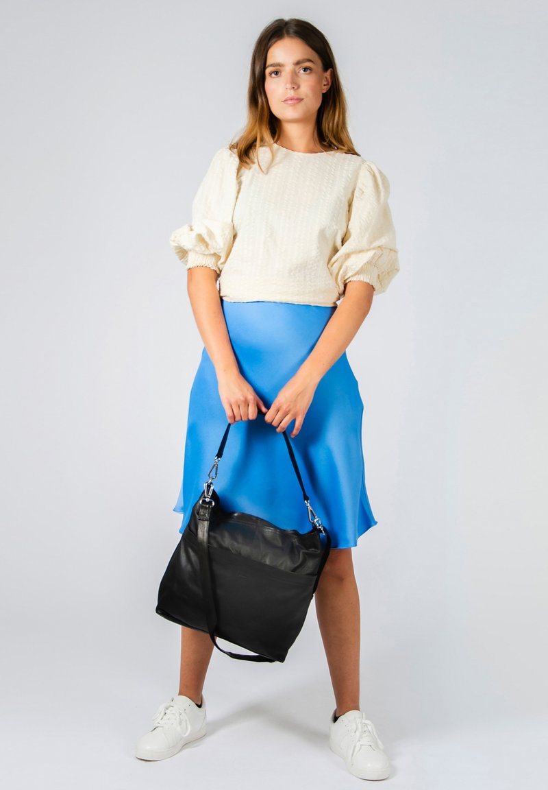 Sac à bandoulière en cuir noir avec une bandoulière réglable, dotée d'une texture lisse et d'un design minimaliste. Le modèle porte un haut crème clair et une jupe en satin bleu.