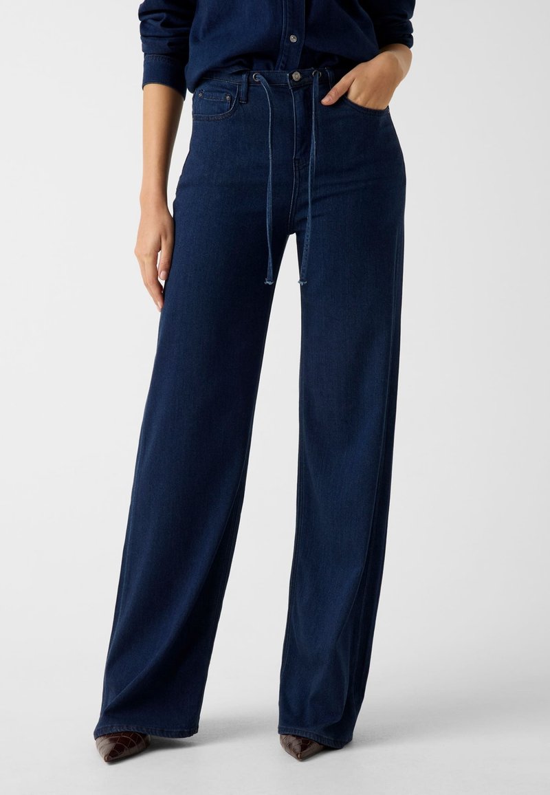 Jean denim foncé à jambe large avec une taille haute, des poches avant et un lien à nouer. Texture lisse et léger éclat, mettant en valeur un design structuré.