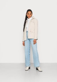 Chaqueta con capucha de color beige claro, con cordones, llevada sobre un jersey blanco de cuello alto, combinada con unos vaqueros de pierna ancha de color azul claro y botas de tobillo blancas con tacón.