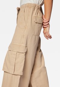 Beige Cargo-Hose aus strukturiertem Material, mit mehreren Seitentaschen, einem elastischen Bund und einer lockeren, entspannten Passform.