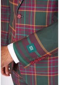 Zelený kostkovaný oblek s červenými a oranžovými akcenty. Obsahuje bílý manžetový knoflík a knoflíková rukáv. Štítek říká "OPPO SUITS." Viditelné detaily texturovan�é tkaniny.