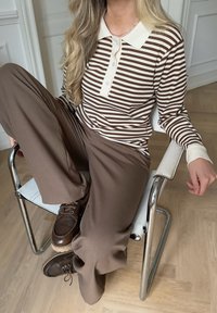 Langärmliges Polo-Shirt mit braun-cremefarbenen Streifen und Knöpfen, kombiniert mit braunen weit geschnittenen Hosen und dunkelbraunen Schnürschuhen, sitzt auf einem Stuhl.