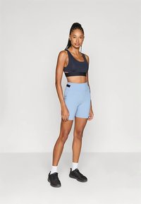 Tumesinise lõikega spordirinnahoidja, millel on lõigetega detail, kombineeritud helesiniste kõrge vöökohaga pükstega, millel on küljetaskud ja mustad aktsendid. Jalga on pandud mustad tossud.