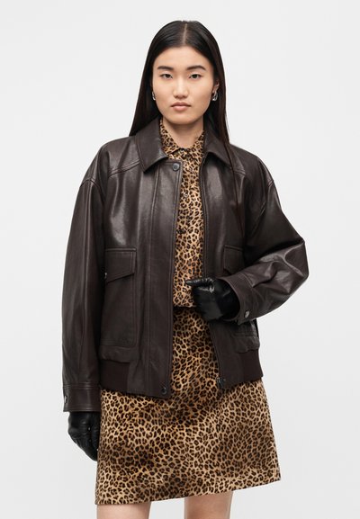 Καφέ δερμάτινο μπουφάν με φαρδύ γιακά, μπροστινές τσέπες και ριμπ τελείωμα, φορεμένο πάνω από φόρεμα με λεοπάρ μοτίβο, συμπληρωμένο με μαύρα γάντια.