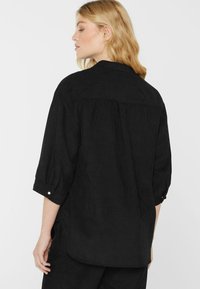 Woman with blonde hair wearing a loose-fitting black corduroy shirt with rolled-up sleeves, shown from the back against a plain background.
