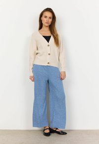 Cardigan bej tricotat cu nasturi aurii, asortat cu pantaloni blue texturat cu picioare largi și pantofi negri, pe un fundal alb simplu.