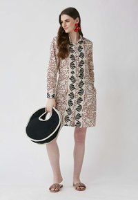 Napapaitin na damit na may butones na gawa sa magaan na tela, na nagtatampok ng abstraktong pulang at itim na mga pattern sa beige na background. Itim na bilog na bag at kulay tans na sandals.