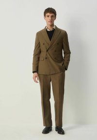 Ruskea kaksirivinen pikkutakki, jossa on kaksi etutaskua, yhdistettynä samanvärisiin housuihin ja mustaan neuleeseen alla. Mustat kengät.