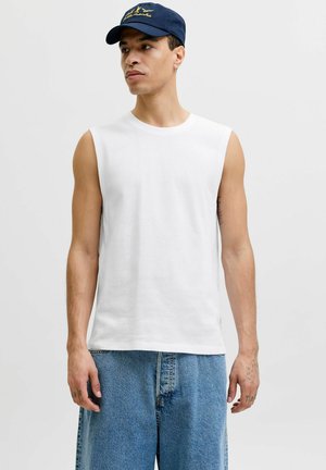 Homme portant un débardeur blanc, un short en jean bleu et une casquette de baseball bleu marine, debout devant un fond clair uni.