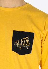 Keltainen puuvillapaidan t-paita, jossa on musta tasku, jossa lukee "SLADE" keltaisella tekstillä ja skeittigrafiikka.