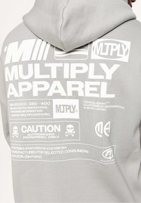 Grå huvtröja av fleecetyg med stora vita texter och grafiska element på baksidan, inklusive logotyper och varningssymboler.