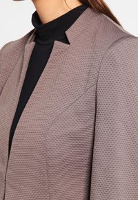 Femme portant un blazer mauve texturé sur un col roulé noir, mettant en valeur le décolleté et les détails des épaules.