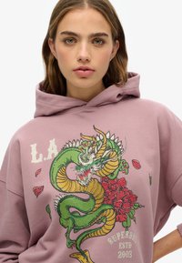 Sudadera de color rosa claro con un gráfico prominente de un dragón en verde y dorado, con acentos florales. Incluye el texto "L.A. Superdry ESTD 2003."