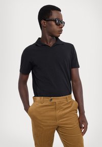 Schwarzes Polohemd mit weicher Textur, kombiniert mit hellbraunen Cordhosen mit Knopfverschluss. Das Modell trägt eine dunkle Sonnenbrille.