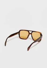 Tortie kvadrātveida saulesbrilles ar dzintara toņu stikliem, platām kājiņām un dubultu tiltu, novietotas uz baltas virsmas.