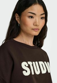 Bruine sweatshirt met "STUDIO" in gestructureerde witte letters. Ronde halslijn en een relaxte pasvorm. Model draagt kleine zilveren ringoorbellen.