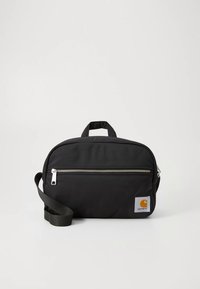 Bolsa de nylon negra con forma rectangular, asa superior y correa ajustable. Cuenta con una cremallera plateada y un parche con el logo en el lateral.