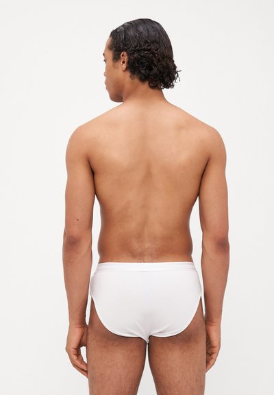 Slip de bain blanc au design ajusté, fabriqué en tissu lisse. Le modèle montre le dos et les épaules nues, avec des cheveux bouclés encadrant le visage.