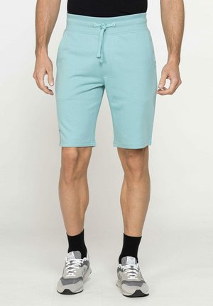 Uomo in piedi che indossa pantaloncini azzurri con coulisse, calzini neri, scarpe da ginnastica grigie e bianche e maglietta nera, su sfondo semplice.