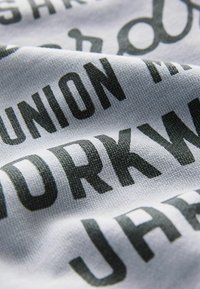 Bílá bavlněná látka s výrazným tmavě šedým typografickým potiskem, obsahující různé slova a fráze uspořádané v překrývajících se vzorech.