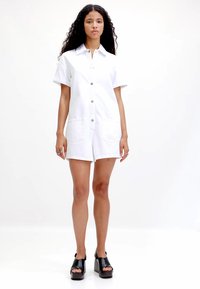 Witte denim korte jumpsuit met korte mouwen, knopen aan de voorkant en twee voorzakken. Model draagt zwarte plateau sandaal. Glad textuur.
