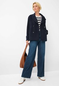 Cappotto doppiopetto blu navy con grandi bottoni, maglietta a righe, ampi jeans in denim blu, sneakers bianche e borsa a tracolla marrone.