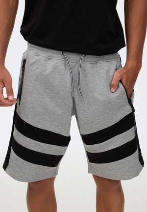 Hombre con pantalones cortos grises con rayas diagonales negras y bolsillos con cremallera, una mano en el bolsillo, camisa negra parcialmente visible.