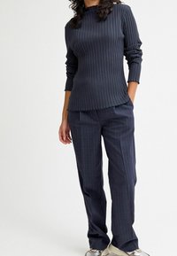 Marineblaue, gerippte Langarmbluse mit hohem Kragen, kombiniert mit karierter marineblauer Hose, die eine lockere Passform aufweist und auf den Schuhen einen subtilen Glanz hat.