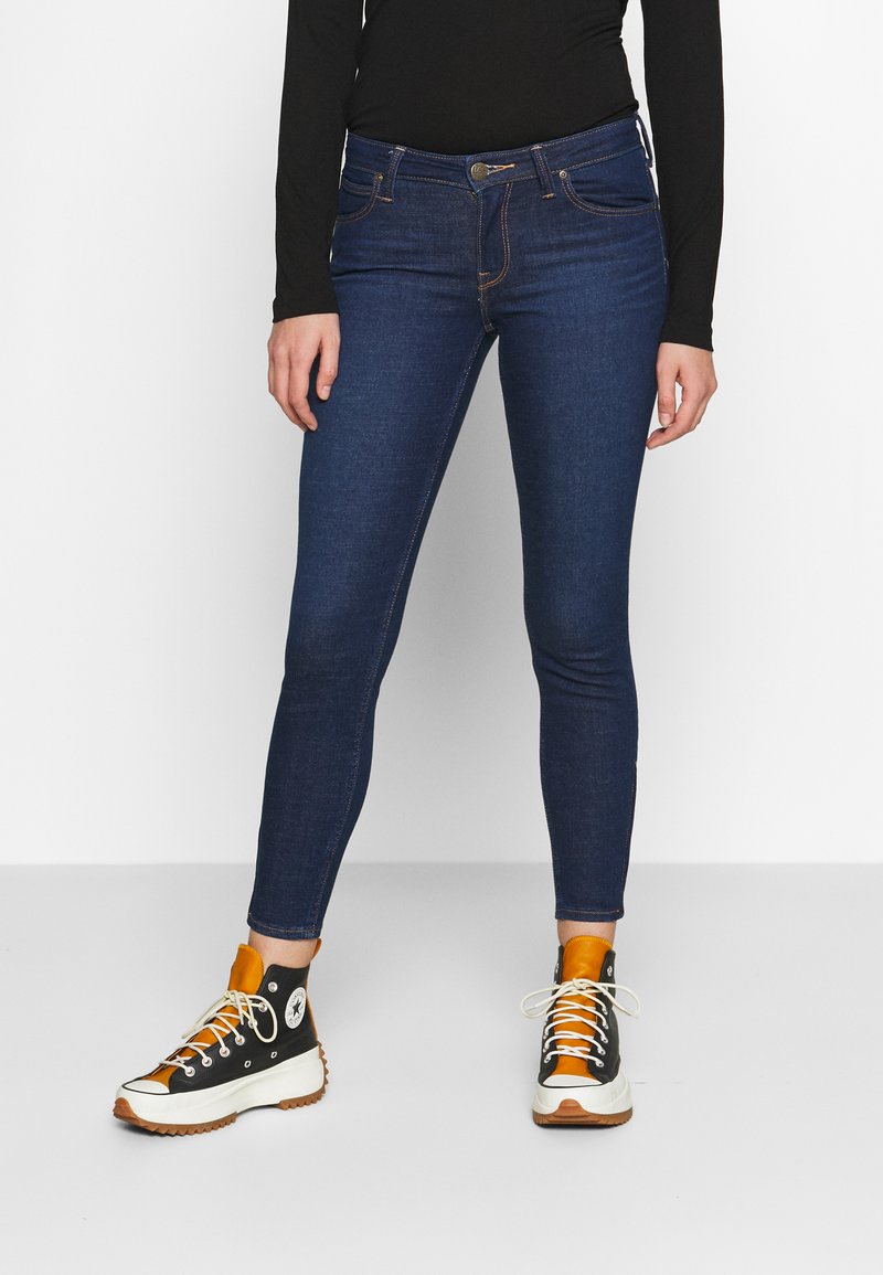 Chude niebieskie dżinsy z kontrastowym przeszyciem i przednimi kieszeniami, w połączeniu z czarno-pomarańczowymi sneakersami typu high-top. Gładka tekstura.