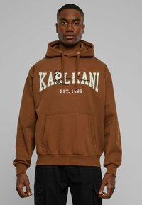 Felpa marrone con coulisse, tasca frontale e testo grafico "KARL KANI EST. 1989" in crema e nero. Materiale in cotone. Design informale.