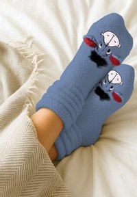 Calze in pile blu con design di Eeyore, caratterizzate da orecchie rosse, un ciuffo nero e una faccia sorridente, poggiate su una leggera coperta.