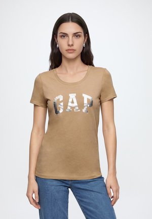 Jeune femme aux longs cheveux foncés portant des boucles d'oreilles en argent, un t-shirt marron GAP avec des lettres métalliques, et un jean bleu, debout sur un fond blanc.