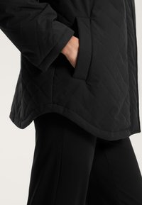 Črna prešita jakna z zaobljenim robom, ki ima horizontalne šive in stranske žepe. Nosi se preko črnih hlač, gladka tekstura.