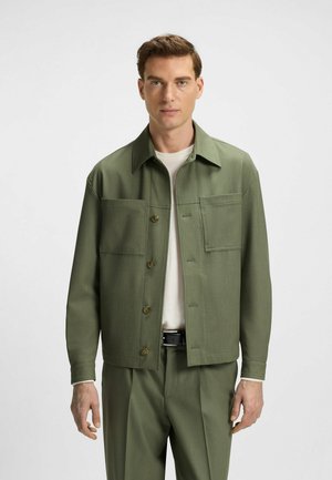 Groene, gestructureerde jas met een platte kraag, twee voorzakken en knoopsluiting, gedragen over een lichtgekleurde shirt en bijpassende broek.