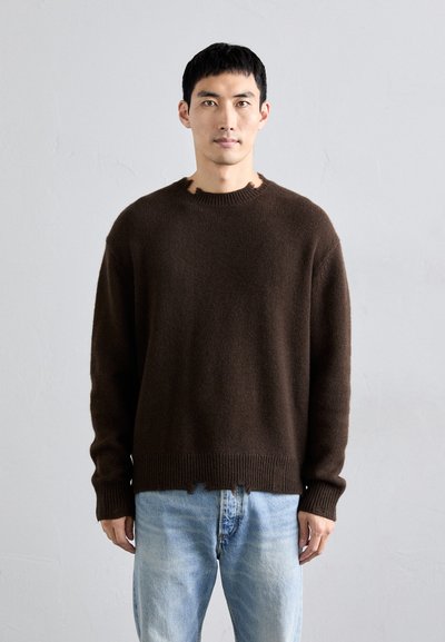 Pull en tricot marron à col rond, avec une finition texturée et un ourlet effiloché. Associé à un jean en denim bleu clair.